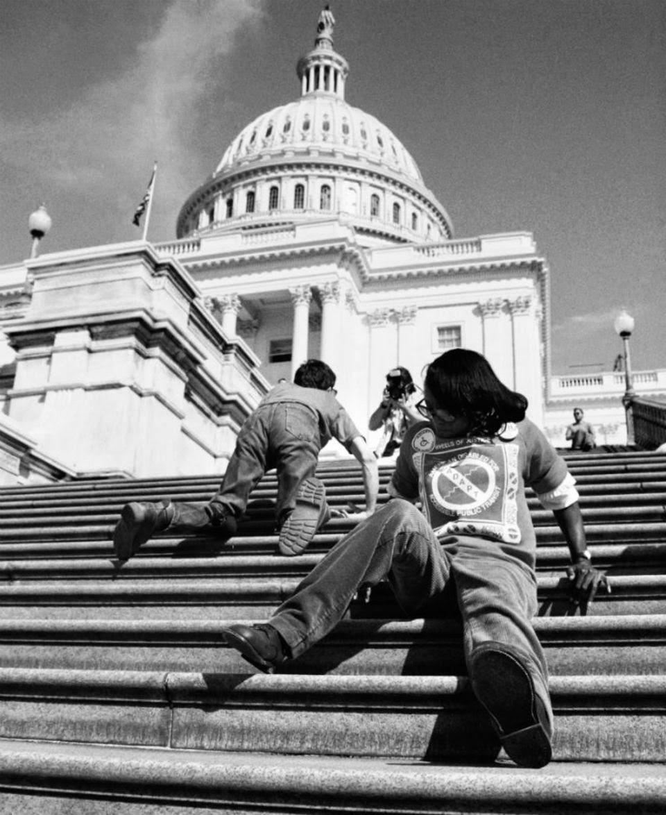 Two young people crawl up the steps outside the US Capitol Building. The image climbs from the bottom right, with the foot of a protester pointing at the camera, all the way to the towering building and white cupola at the top left.